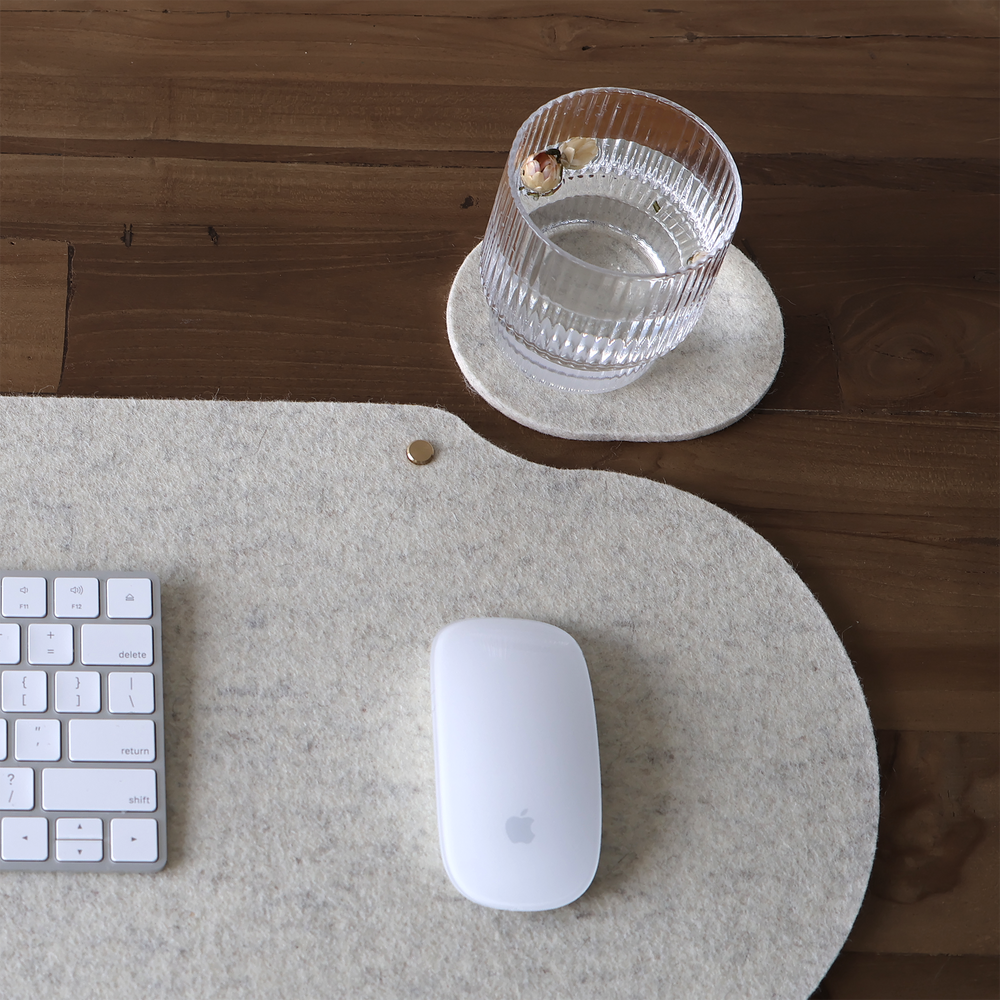 Natural wool felt desk mat with white keyboard and mouse on a wooden desk, beside a matching felt coaster and glass.