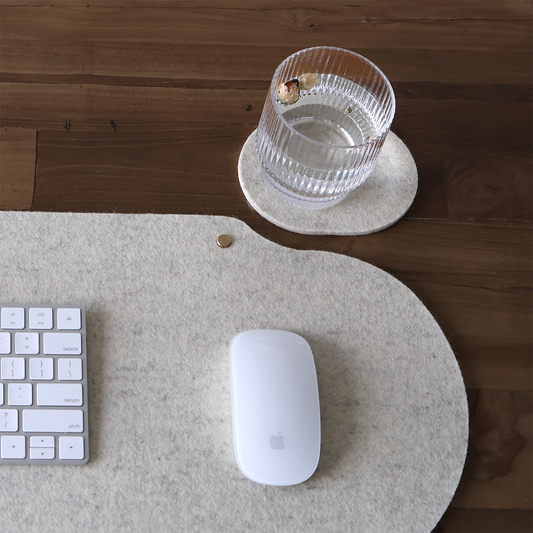 Natural wool felt desk mat with white keyboard and mouse on a wooden desk, beside a matching felt coaster and glass.